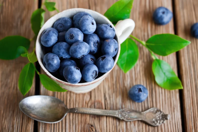 Blueberries in a white ceramic cup with green leaves and a spoon on a rustic wooden table