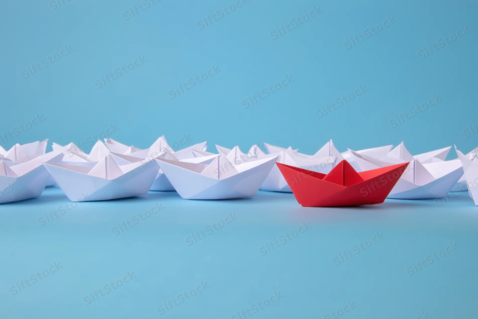 Red paper boat leading a fleet of white boats on a blue surface