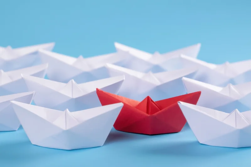 Red paper boat among white boats on blue background