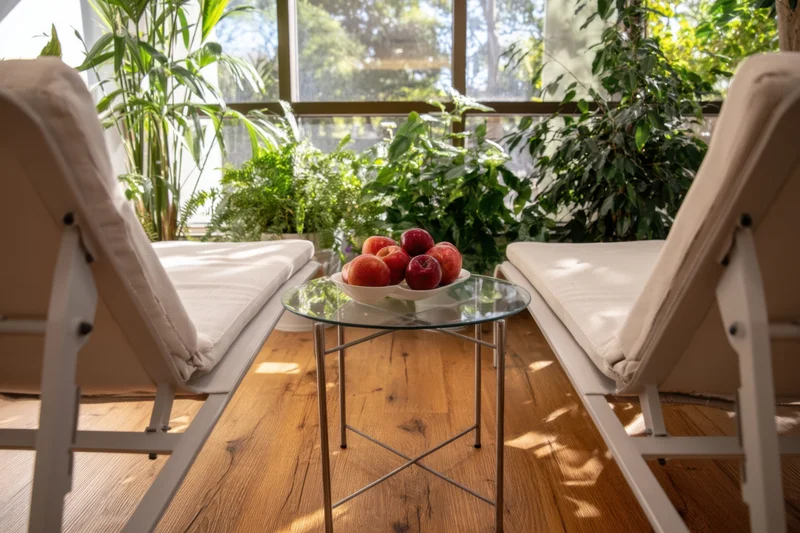 Two lounge chairs with a glass table holding a bowl of apples in a sunlit room with plants and large windows