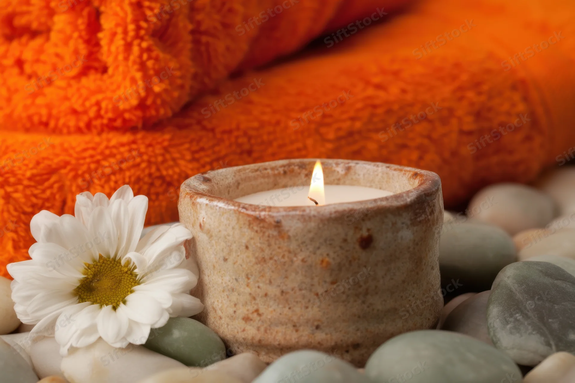Lit candle in ceramic holder surrounded by smooth stones and a white daisy near an orange towel in a spa setting