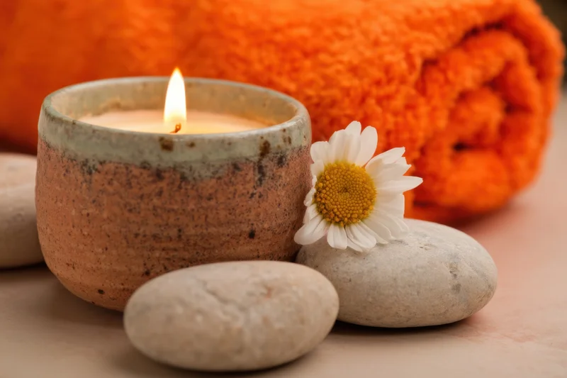 Lit candle in ceramic pot beside smooth stones and daisy flower with orange towel in background