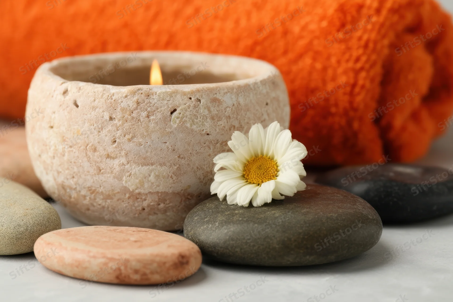 Lit candle in textured ceramic holder with daisy and smooth stones beside orange towel on light surface