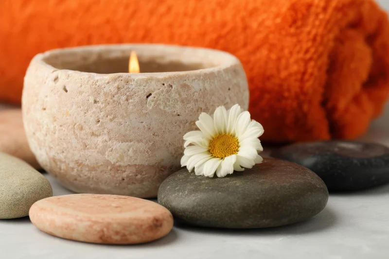 Lit candle in textured ceramic holder with daisy and smooth stones beside orange towel on light surface