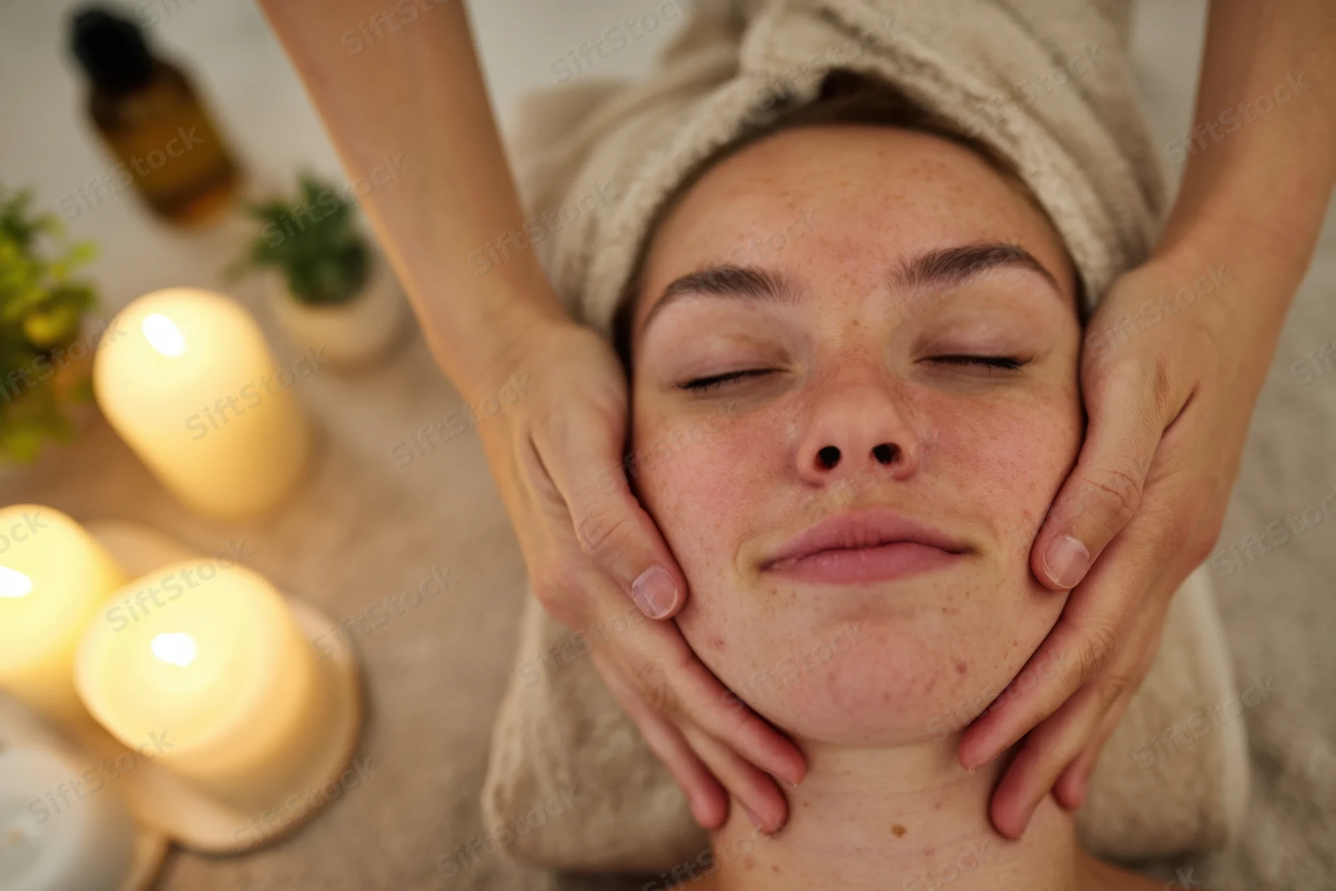 Woman receiving facial massage in spa setting with candles and towel on head