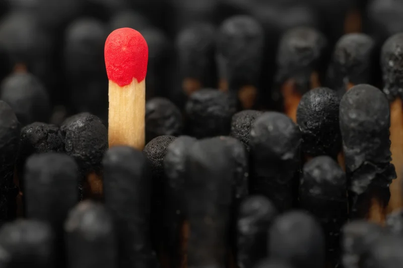 Single red-headed matchstick standing upright among burnt black matchsticks in close-up view