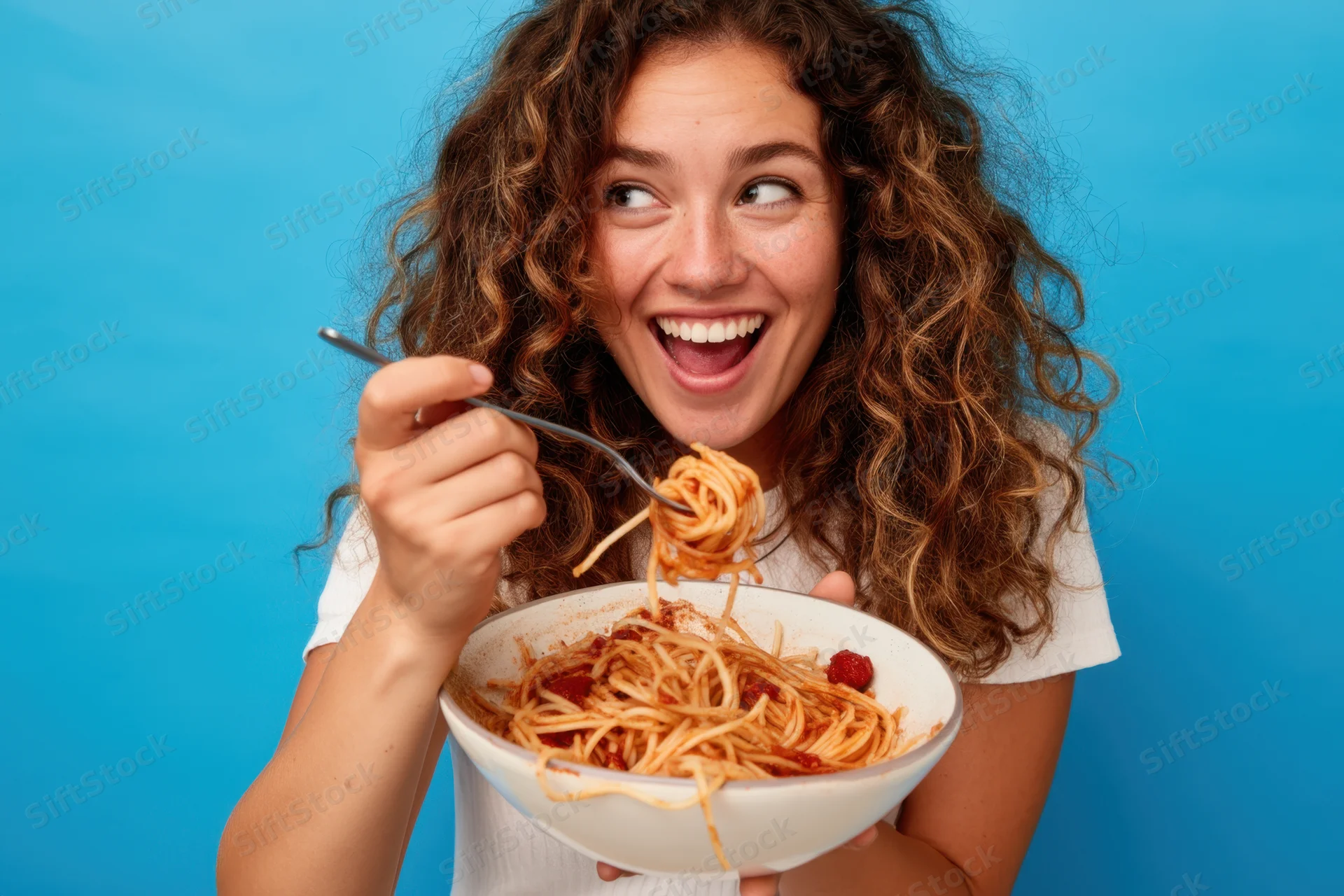 Woman laughing while eating spaghetti from a bowl with a fork against a blue background