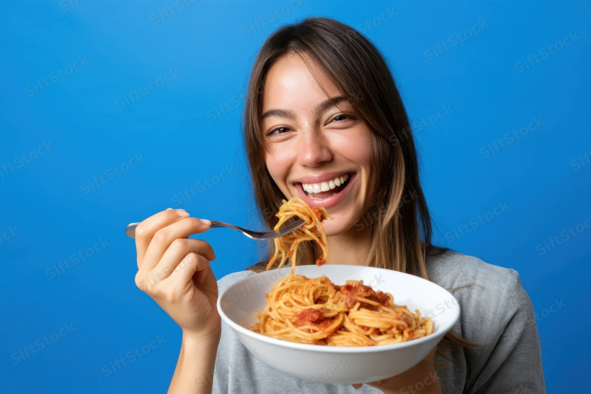 Smiling woman eating spaghetti from a white bowl against blue background