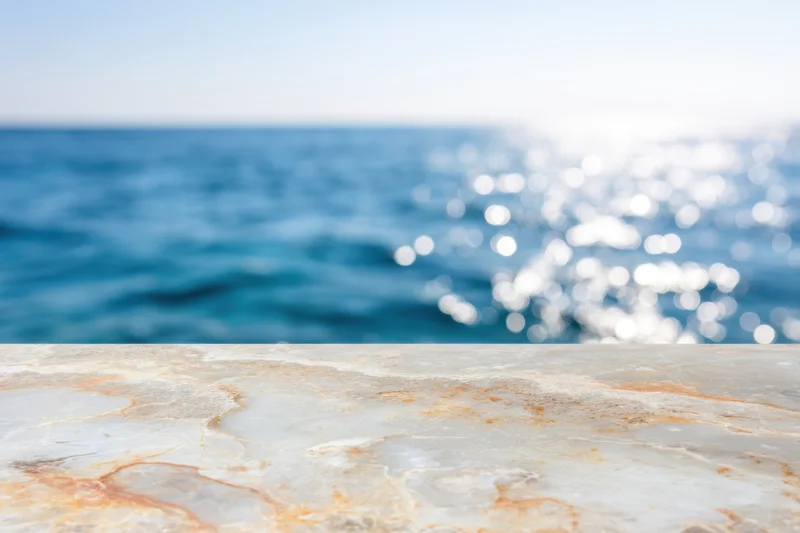 Smooth marble tabletop in foreground with blurred ocean and sunlight in background