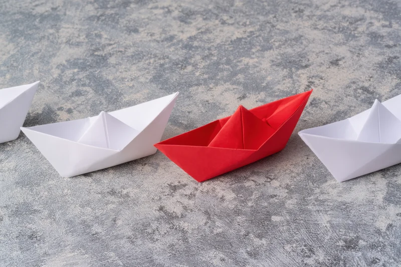 Red origami boat positioned among white boats on a gray textured surface