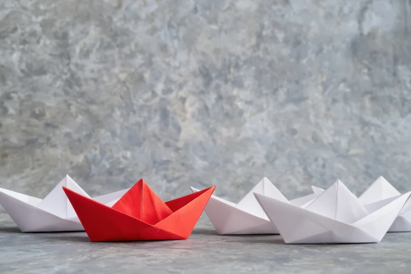 Red origami boat leading a group of white boats on a textured surface