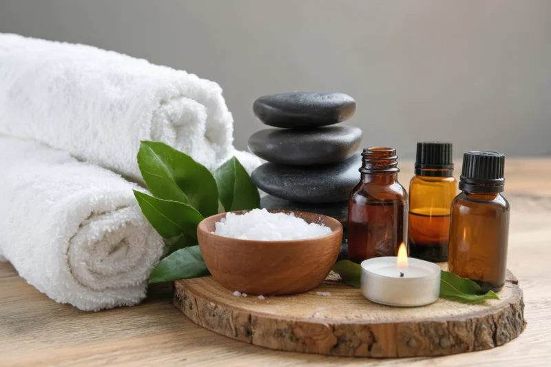 Stack of smooth black stones and essential oil bottles with lit candle and bath salt on wooden tray beside rolled white towels
