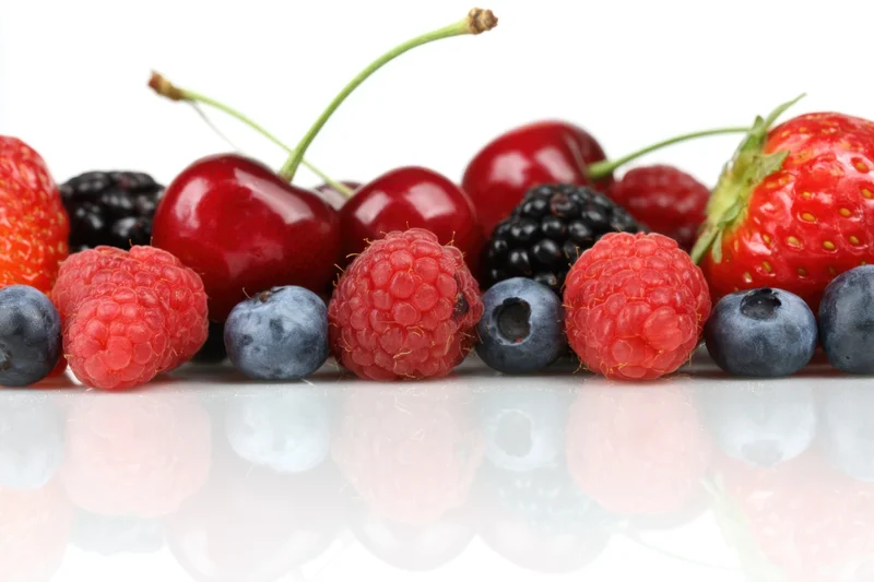 A row of fresh berries including strawberries, raspberries, cherries, blueberries and blackberries arranged on a reflective white surface