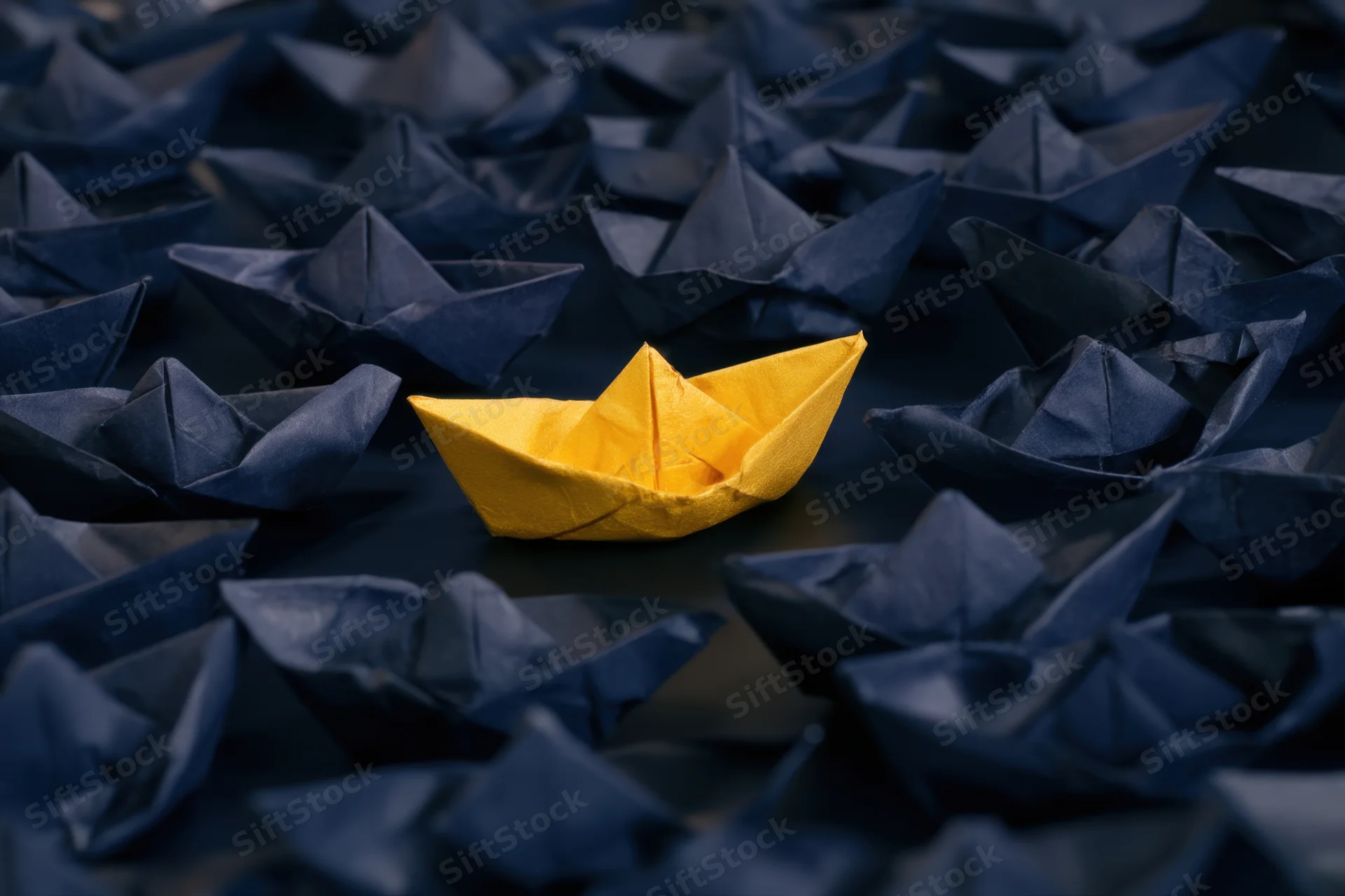 A yellow paper boat centered among navy blue paper boats on a dark surface
