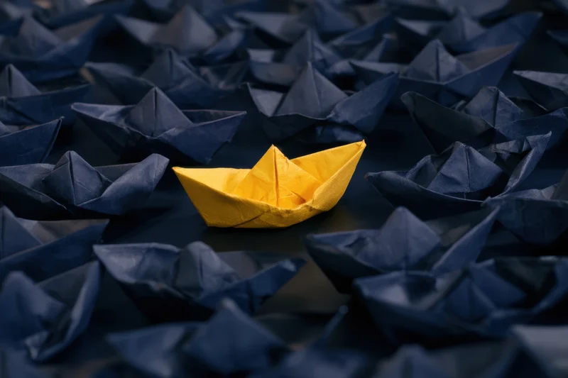 A yellow paper boat centered among navy blue paper boats on a dark surface