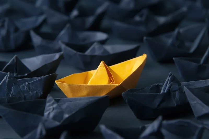 A gold paper boat standing among navy blue paper boats on a dark surface