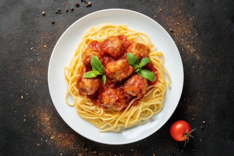 Spaghetti topped with meatballs in tomato sauce on a white plate with basil garnish on dark surface