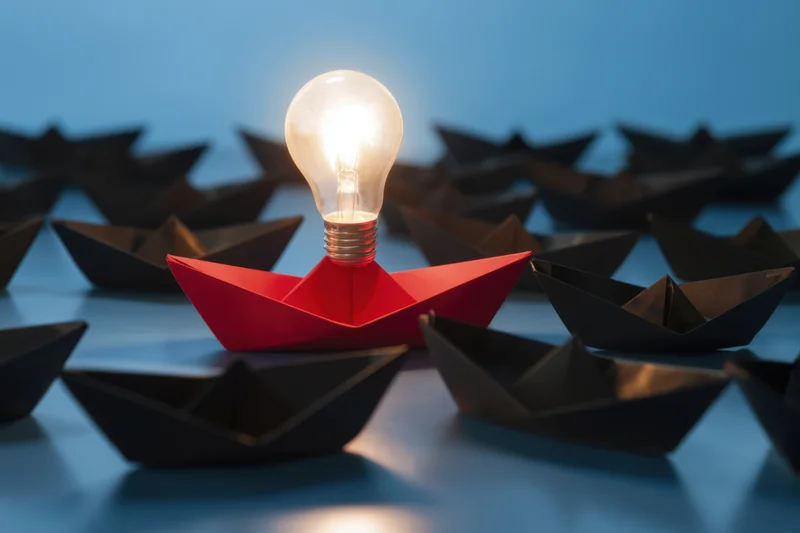 Red paper boat with glowing lightbulb among black boats on blue surface