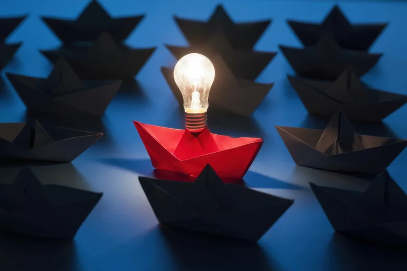 Red paper boat carrying a glowing lightbulb among dark boats on blue surface