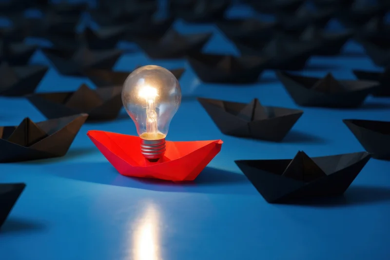 A red paper boat carrying a glowing lightbulb among black boats on a blue surface