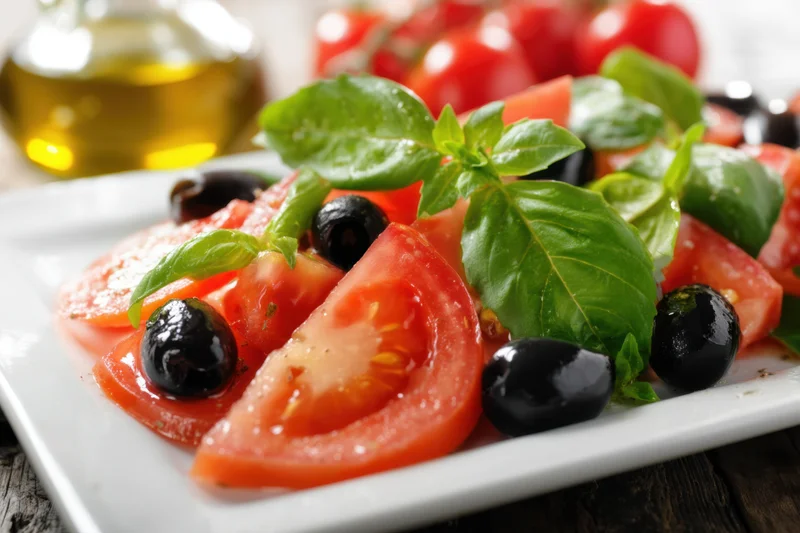 Sliced tomatoes with fresh basil and black olives arranged on a white plate with olive oil in background