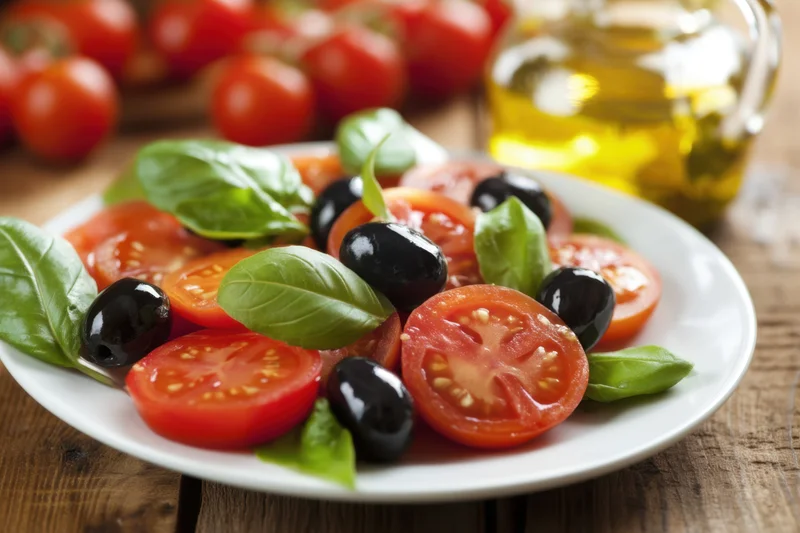 Sliced tomatoes and black olives with fresh basil leaves arranged on a white plate on a wooden table