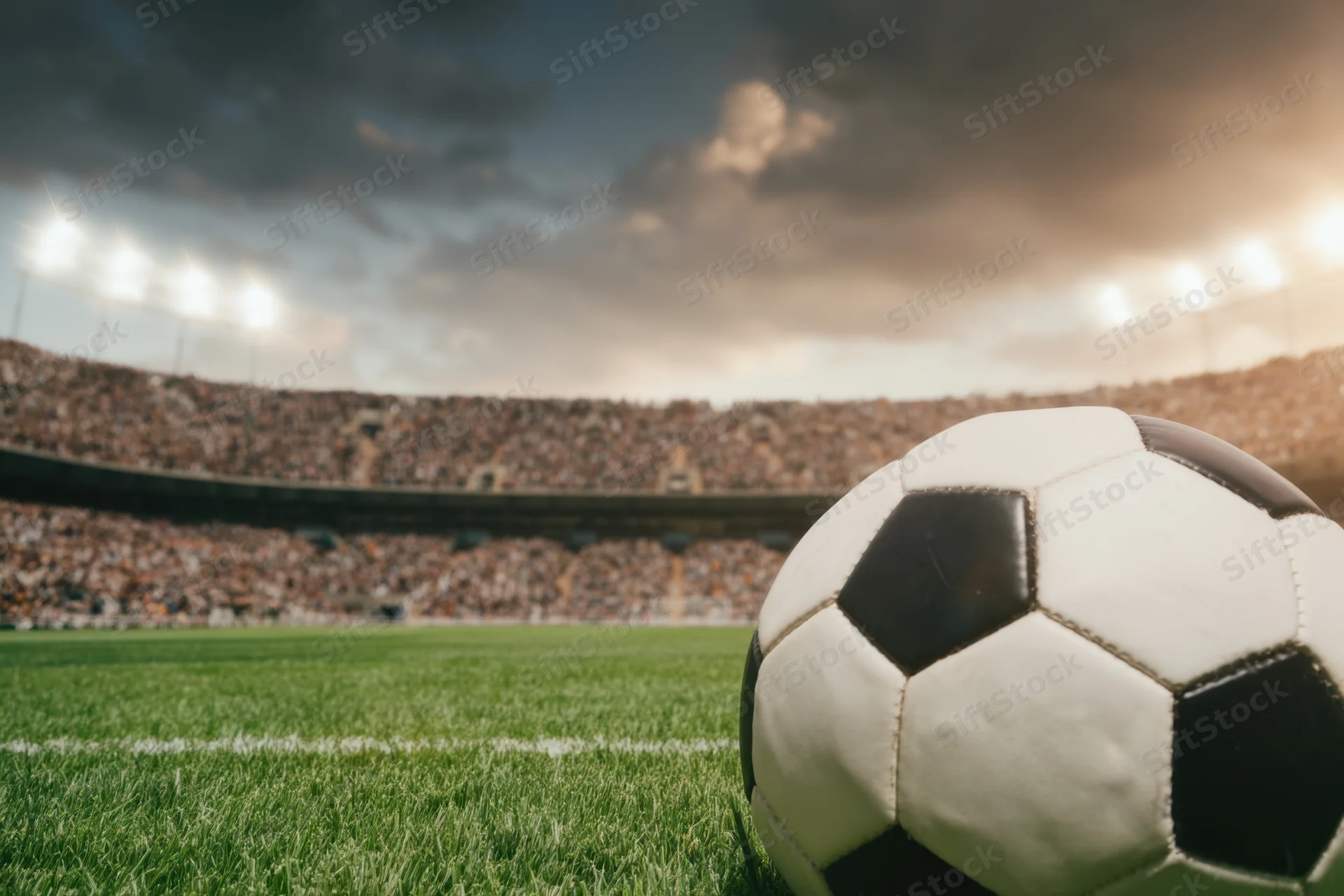 Soccer ball resting on grass field in front of crowded stadium under floodlights at dusk