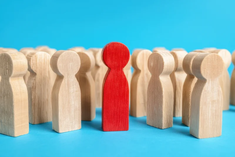 Red wooden figure standing among a group of beige figures on a blue background