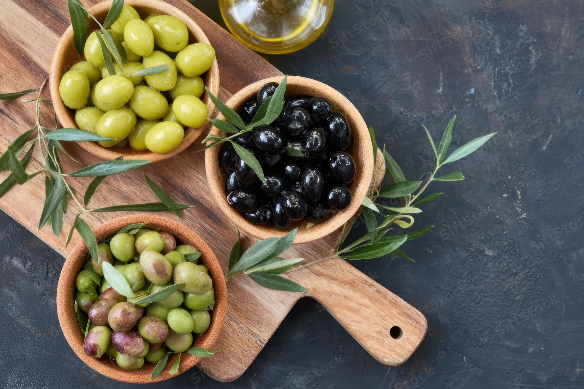 Wooden serving board with bowls of green, black and purple olives arranged with olive leaves and olive oil bottle on dark surface