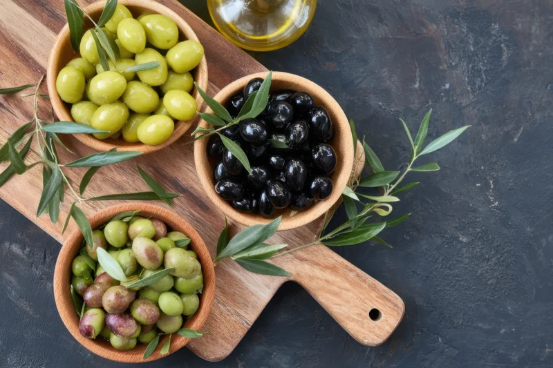 Wooden serving board with bowls of green, black and purple olives arranged with olive leaves and olive oil bottle on dark surface