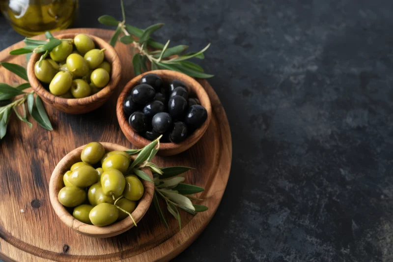 Wooden serving board with bowls of green and black olives arranged with olive branches and oil on dark surface