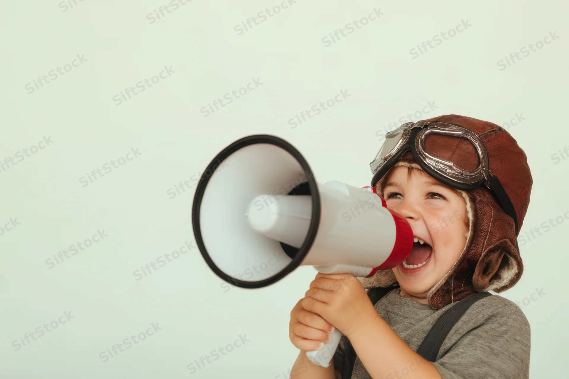 Young boy wearing vintage aviator hat and goggles shouting into a megaphone indoors