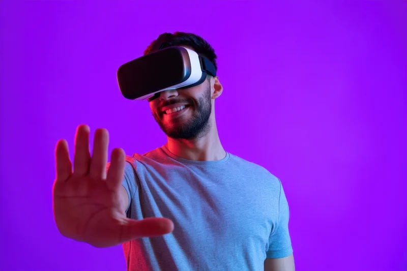 Young man wearing virtual reality headset smiling and gesturing with hand against purple background
