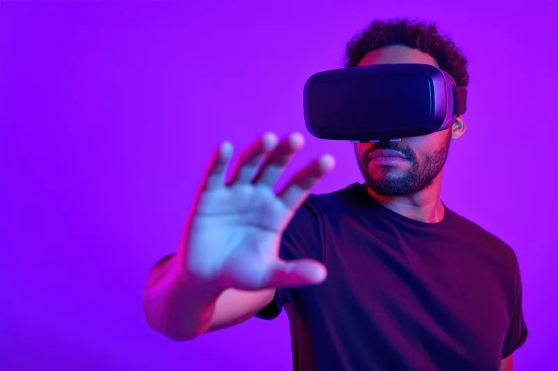 Young black man wearing virtual reality headset reaching forward with hand in neon-lit studio