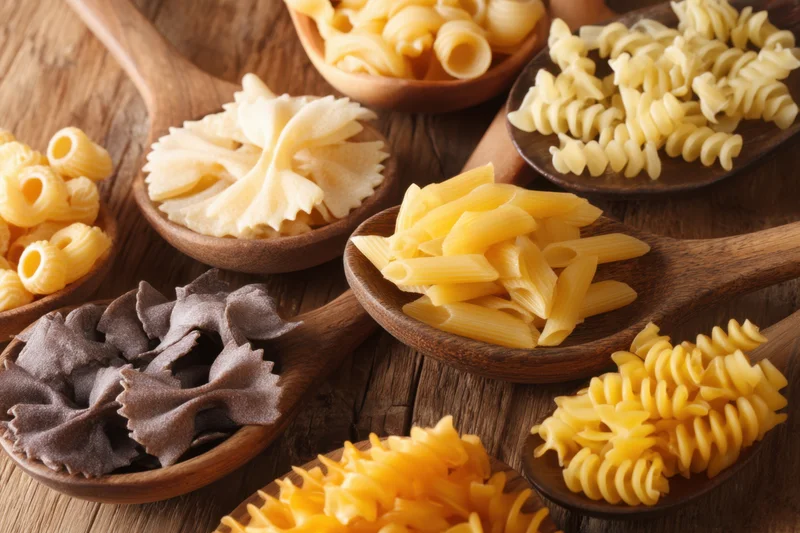 Various uncooked pasta types arranged on wooden spoons on a rustic table
