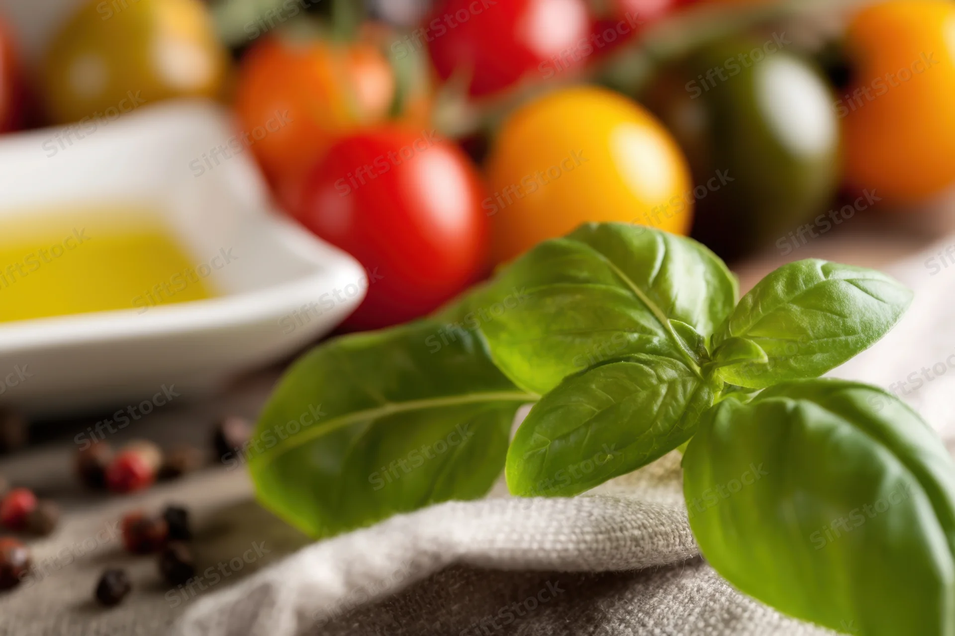 Fresh basil leaves in focus with colorful cherry tomatoes and peppercorns on a linen cloth in a kitchen setting