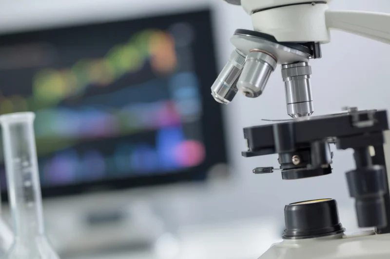 Close-up of a microscope in a laboratory with blurred monitor displaying data in background