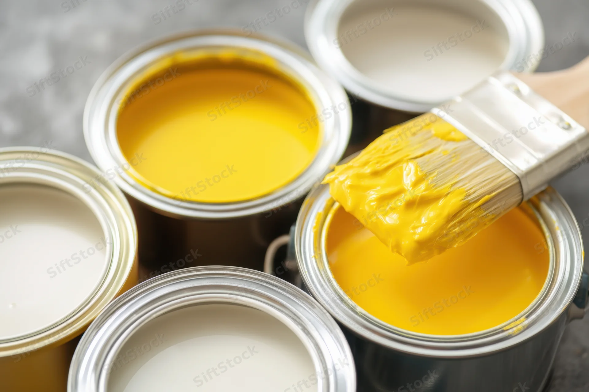 Paintbrush loaded with yellow paint hovering over open paint cans in a minimalist setting