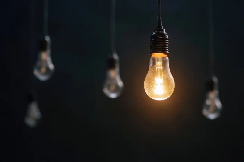 Single illuminated lightbulb hanging among unlit bulbs against dark background