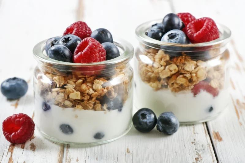 Two glass jars filled with layered yogurt, granola and mixed berries on a rustic wooden surface