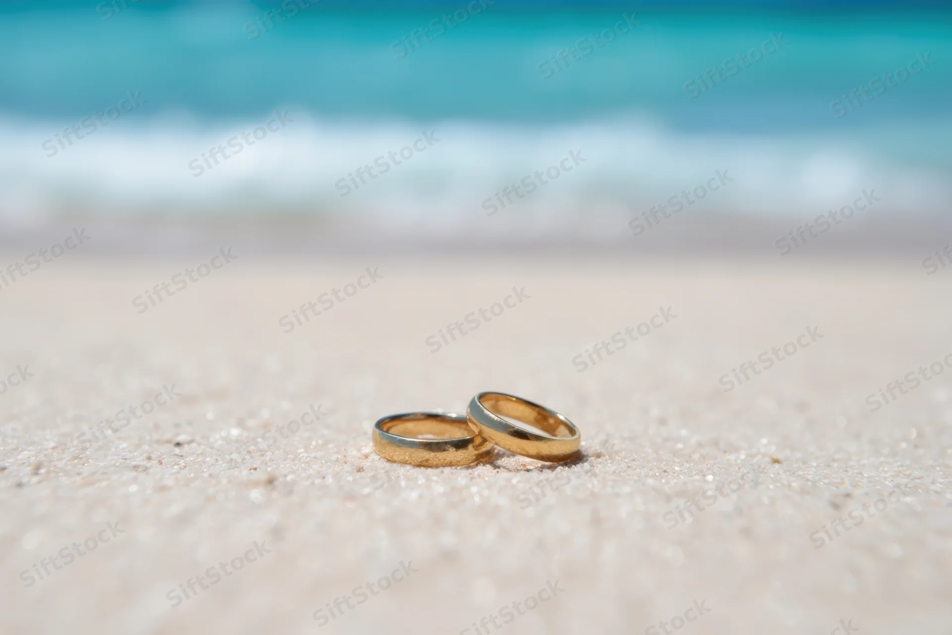 Two golden wedding rings resting on sandy beach near ocean waves