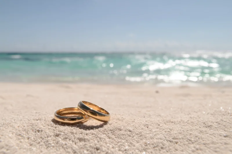 Two golden wedding rings resting on sandy beach near ocean waves
