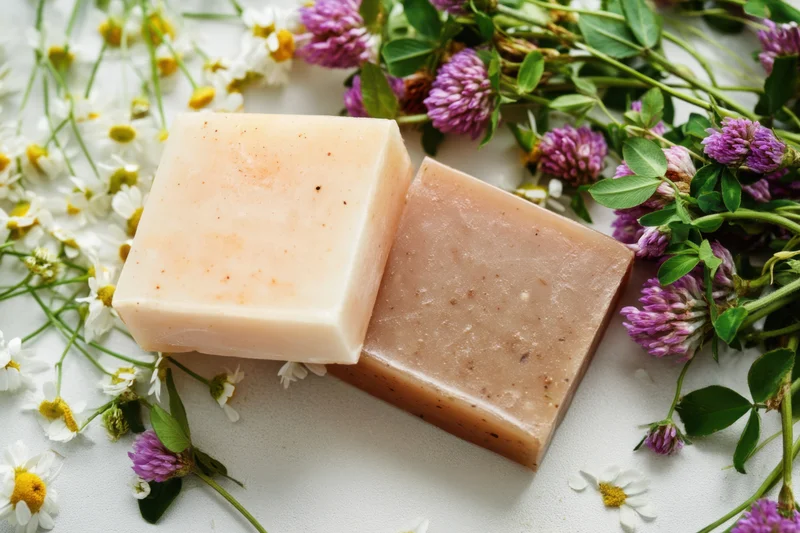 Two natural soap bars resting on a light surface surrounded by clover and chamomile flowers