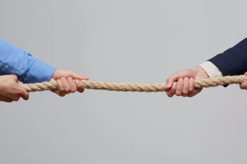 Two pairs of hands gripping a rope in a tug-of-war against a plain gray background