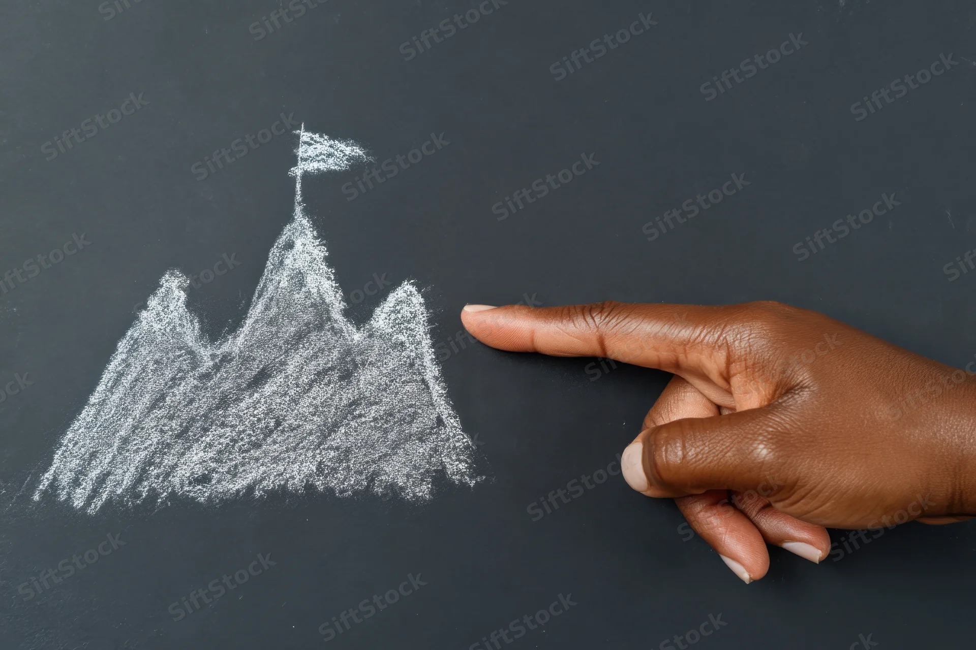 Hand pointing to chalk-drawn mountain with flag on summit on blackboard