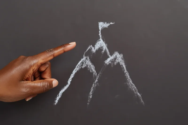 Hand pointing to chalk drawing of mountain peak with flag on blackboard