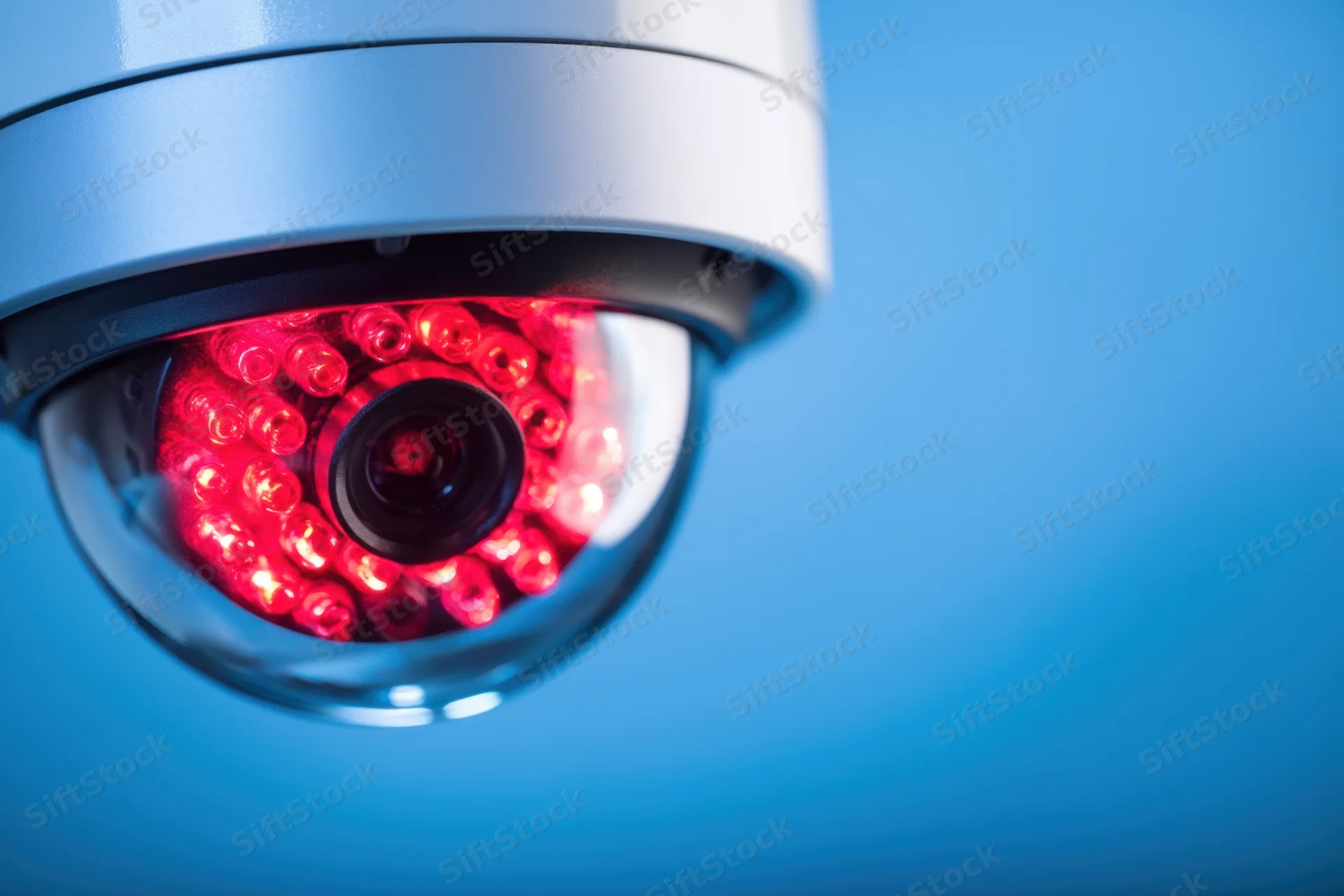Close-up of a silver dome cctv camera with red led lights glowing against a blue background