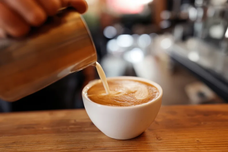Barista pouring steamed milk into a cup of coffee on a wooden counter in a cafe