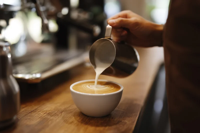 Barista pouring steamed milk into a cup of coffee on a wooden counter in a cafe