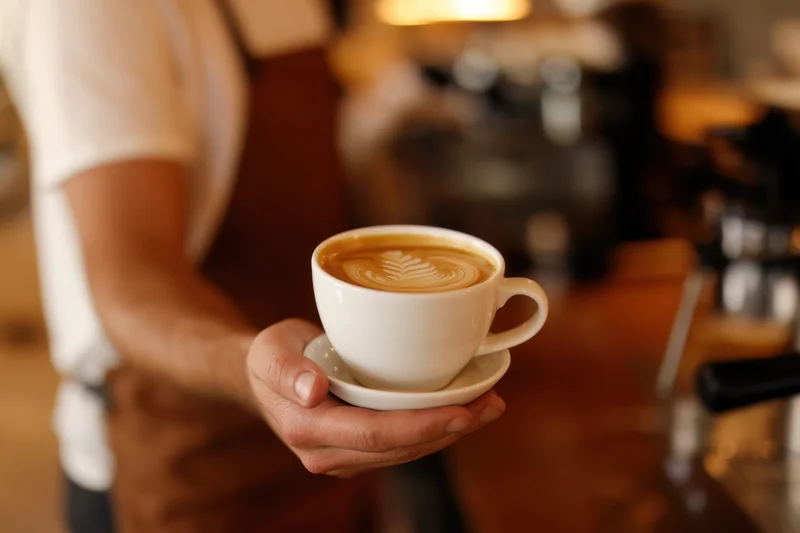 Barista holding a latte art coffee in a white ceramic cup at a cafe counter